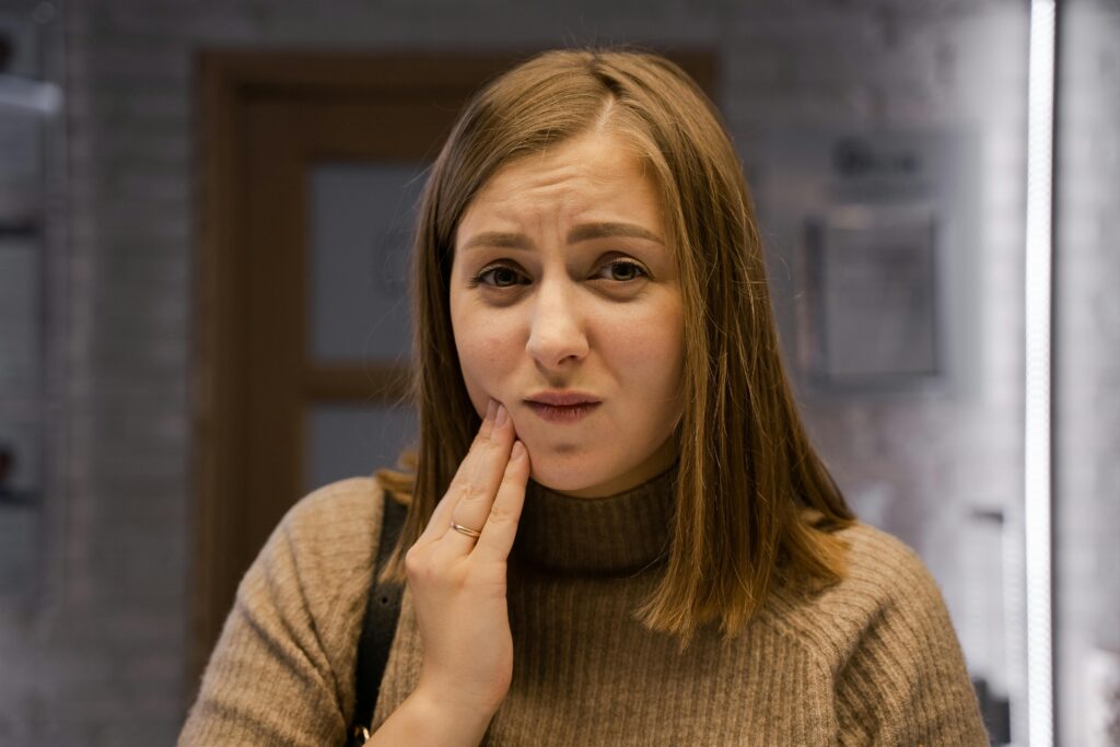 Caucasian woman expressing toothache discomfort, wearing a brown sweater indoors.