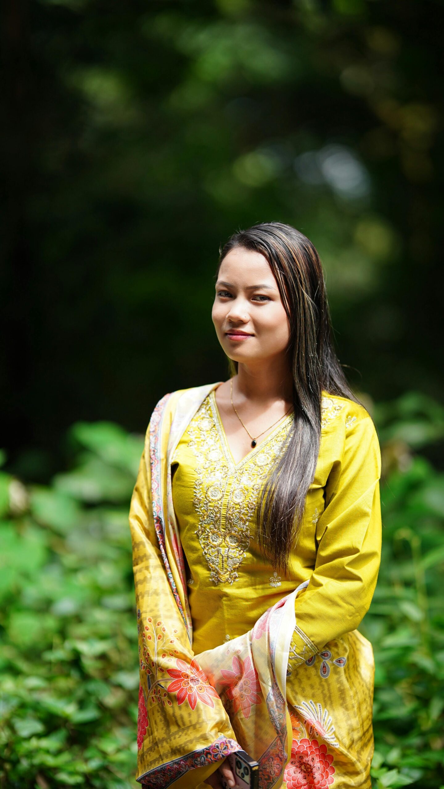 Young woman in traditional yellow attire smiling outdoors, surrounded by greenery.
